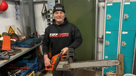 Clinton Perry is in a workshop smiling broadly as he wields a chainsaw. He is wearing a black woolly hat and black sweatshirt with branding reading: Tough Stuff. There are lockers behind him and a workbench full of kit to his right.
