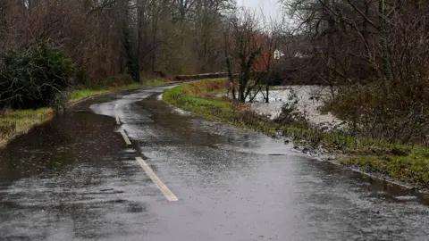 Pacemaker A flooded country road with standing water and nearby river overflowing into surrounding trees.