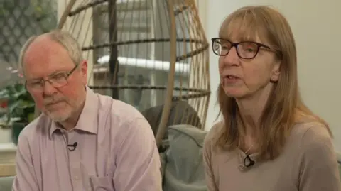 Emma, a woman with long fair hair and dark-rimmed glasses and Geoff, a man with grey hair and a trimmed, grey beard, sitting on a sofa in a living room.