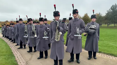 A row of soldiers in long grey coats and tall black fur hats with black feathers holding a variety of silver brass instruments on a footpath with grass on either side.