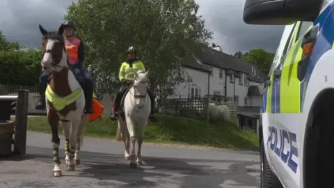 Two horses are seen approaching the camera with riders wearing high-viz jackets and a police car seen to the right. 
