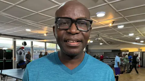 Nicola Haseler/BBC Tony Egynjobi standing in a room, with men playing table tennis behind him. He has on black glasses and a blue T-shirt. There are lights in the ceiling. 