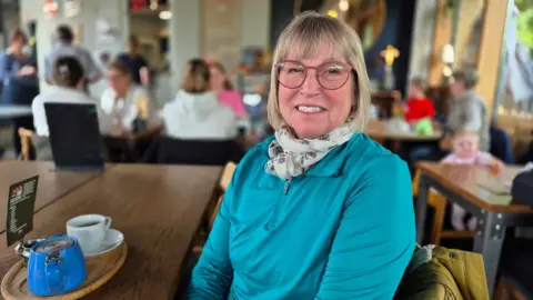 Stuart Woodward/BBC Anne Nicol sits in a cafe with a white cup and a blue teapot next to her on a table. She is wearing a turquoise quarter-zip top with a light-coloured scarf around her neck. She has round spectacles, blonde hair and is smiling at the camera.