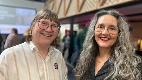 Kate Wyatt and Marina Strinkovsky stand next to each other smiling. Both wear glasses. Kate wears a white shirt with thin pale stripes. Marina wears a polka dot jacket and has long hair.