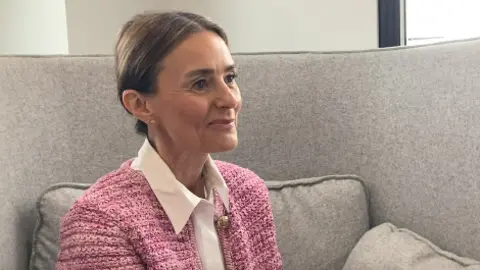 BBC Leyla Yildirim is sitting on a grey sofa. She is wearing a pink blazer and a pink shirt. She has her brown hair neatly tied back and she is smiling.