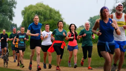 Runners in a park following each other on a gravel path. Many wear T-shirts and shorts. Greenery and trees can be seen behind them.