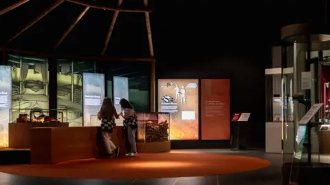 Milton Keynes Museum A dark room with an circle orange carpet and screens along the walls. There are two young girls looking inside a glass casing - situated in front of the screens. It is part of the new gallery. 