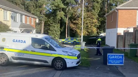 A police car parked in a street with terraced houses and trees in the background. A police officer stands behind white police tape which seals off the area.