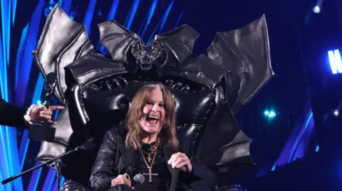 Getty Images The image shows two men. Jack Black is standing up. He is wearing a dark suit with a white shirt. He has shoulder-length brown hair and a beard. The second, Ozzy Osbourne, is still in large black leather bat-shaped throne. He is smiling. He had shoulder-length brown hair long dark clothes and a crucifix around his neck. They are in front of a blue and black backdrop. 