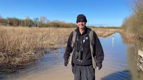 Pete is stood in a waterway. He wears a gloves and a hat and carries a pair of binoculars around his neck. The reed bed is behind him.