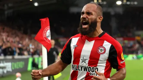 Bryan Mbeumo in red and white striped Brentford shirt shouts and punches the air in front of him with this right hand while celebrating next to a corner flag with a re Brentford flag on top of it