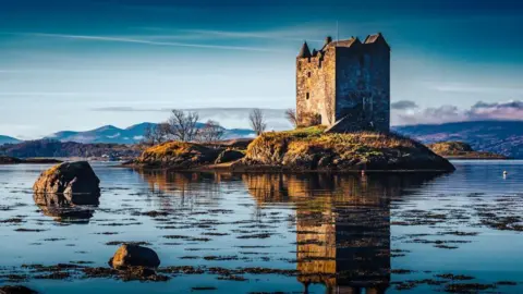 Robert Westerman A historic stone castle sits on a small grassy island surrounded by calm water, reflecting the structure and nearby rocks. The scene is framed by distant mountains under a deep blue sky with light clouds.