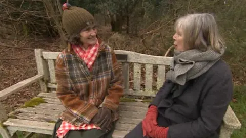 Two women, wrapped up in coats and gloves, smile as they sit next to each other on a park bench. The weather outside is cold and there is no leaves on the trees.