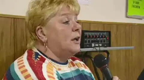 Haney's screen door to a public meeting. Microphone holds. Short blond hair and a large golden pulley earrings. He wears a very colorful jumper