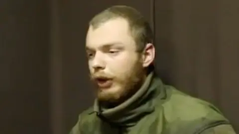 A man believed to be James Scott Rhys Anderson, 22, a former British Army soldier captured while fighting for Ukraine, is seen wearing green military clothing and sat in front of a plain brown background.