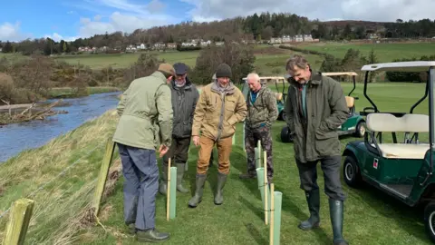 Steven Bridgett A group of men standing by a river edge with golf buggies next to them 