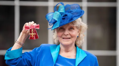 Jo Hamilton holds up her OBE medal which is adorned with red ribbons. She has curly, shoulder-length, blonde hair and wears a bright blue outfit including an ornate, ribboned hat, and a matching jacket and blouse.