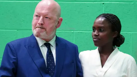 Reform UK candidate Simon Danczuk and his wife Claudine Uwamahoro at the Rochdale by-election count hall in February 2024 at Rochdale Leisure Centre. He is wearing a navy blue suit, she wears a white top.