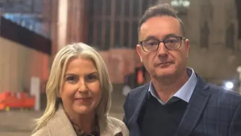 BBC Christine wears a beige coat and has blonde hair, she stands next to Richard who wears a navy checked blazer and navy jumper with light blue shirt. He wear glasses and has short dark hair.