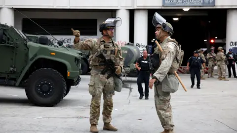 Two National Guard soldiers, wearing camouflage uniforms and helmets with visors lifted up - one pointing with the little of his right hand - stationed at the Metropolitan Detention Center, MDC, in Los Angeles, with more soldiers and police in the background.