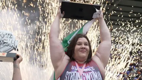 Rebecca Roberts lifting up a trophy and a Welsh flag with fireworks going on behind her