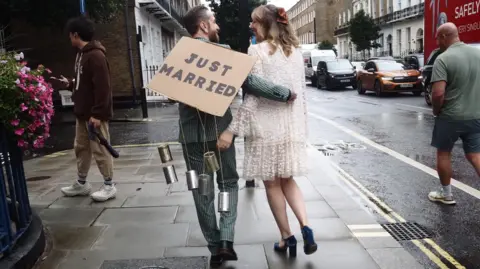 HANDOUT Chris and Sam Jamieson-Green walk along the street. Sam is wearing a white wedding dress and has her arm around the back of her new husband Chris who is wearing a green and white chequered suit with a cardboard sign saying 'just married' on his back.