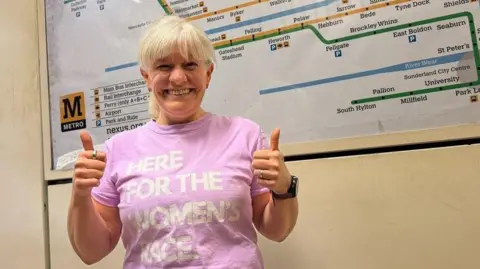 Nexus A smiling Alice Townend with blonde hair is standing in front of a large Tyne and Wear Metro map giving two thumbs up. She is wearing a pink t-shirt with the slogan 'HERE FOR THE WOMEN'S RACE' in white lettering. The setting is a Metro station with Nexus Metro branding behind.