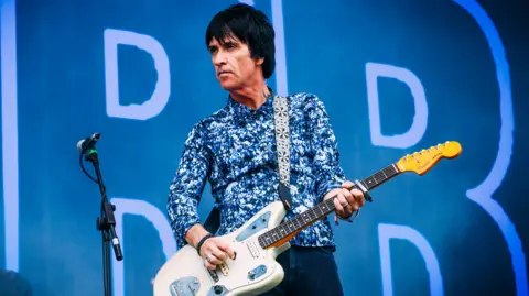 BBC Johnny Marr, with short black hair and wearing a blue, white and black speckled shirt and blue jeans, plays a white guitar next to a microphone