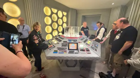 Several people stand around a 1983 TARDIS Console Room replica. In the middle is a round console space which is grey with sharp edges. It has buttons all over it of different sizes and a clear cube in the middle with squares inside it. The walls are grey with yellow circles on it.