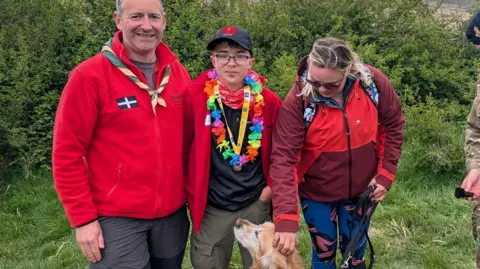 Carrick Deg Karn Explorer Scouts James, Alfie and Melanie stood together with their dog. Melanie is looking down and stroking the golden retriever. James and Alfie are wearing red fleeces with the Cornish flag on the left. 