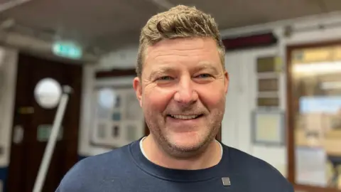 Damien Bolton is smiling broadly at the camera. He has short brown hair and blue eyes and is wearing a blue sweatshirt. There are frames hanging on the wall behind him.
