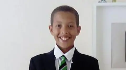 Noah smiling at the camera as he stands against a white wall. He is wearing his school uniform which is a black blazer, green and black striped tie and a white shirt. He has short black hair.