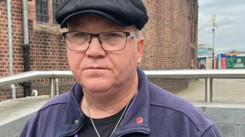 Jonny Humphries/BBC Graham Rudge, who has glasses, is wearing a black flat-cap and a purple fleece, looks into the camera with a serious expression. 
