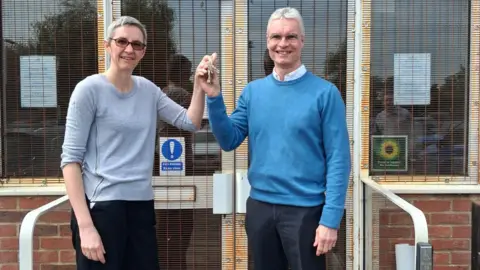 Two people standing outside, holding a set of keys between them. The woman, to the left has on a grey jumper, dark trousers and has short grey hair and glasses. The man has on a blue jumper, shirt and dark trousers. He also has glasses on and short grey hair. 