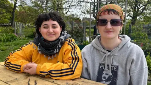 Misha, left, wearing a yellow jacket with three black stripes down the arm. She has short dark hair and is wearing a scarf. Aoibh, right, wearing sunglasses and a green and orange floral bandana. She is wearing a grey hooded jumper with an image of James Connolly on the front.