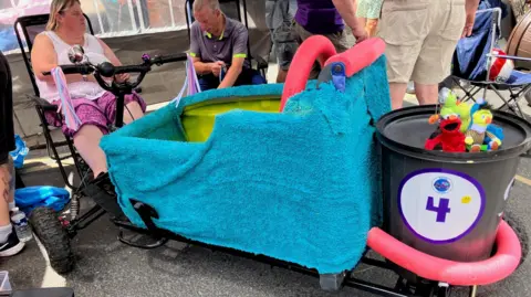 A participant prepares to drive a homemade soapbox cart designed to look like a bathtub during an outdoor event. The cart is decorated with a blue towel, pink pool noodles, a black bucket displaying the number 4, and small soft toys attached to the front.