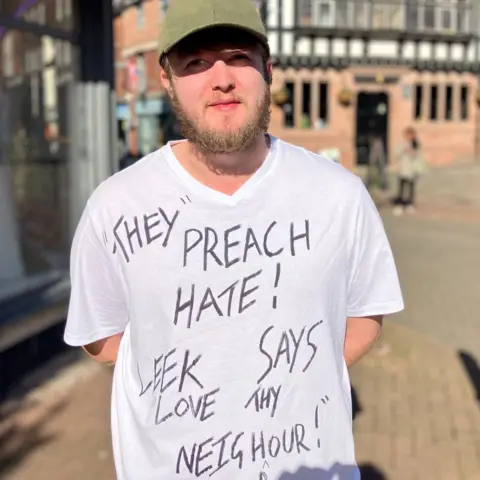 A man stood in Leek town centre looking at the camera with a t-shirt on that reads: "They preach hate! Leek says love thy neighbour".