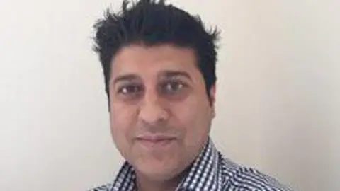 Jonathan Fine Sheraz Ahmed is staring into camera in a tightly framed shot. He has dark hair and yes and a blue and white checked shirt.