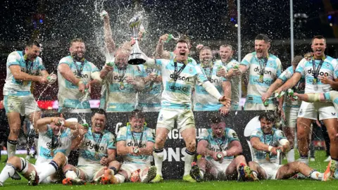Bath players celebrate with the trophy and spray champagne in front of fans