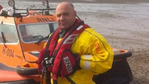 A man with a red floatation vest and a yellow jacket standing in front of an orange boat on the coast. On it is the word RNLI.