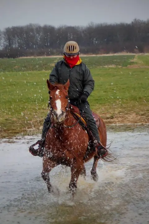 Julie Doorne Julie Doorne rides her brown horse through a body of water, she is wearing a black jacket, red scarf, and riding hat.