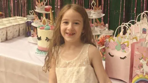 A little girl is standing in a village hall in front of a decorated table filled with party bags, a unicorn-themed birthday cake and smaller unicorn cakes on a cake stand. The girl is wearing a long white dress and is holding a light blue balloon. There is another table filled with presents in the background.