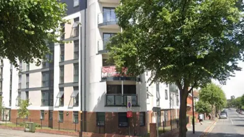 Manor Park Court student accommodation in Coventry is a block of flats in a white modern building with trees outside. There are signs on a balcony and some of the windows are open.