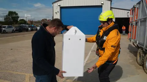 Shaun Whitmore/BBC A man in a life vest and yellow jacket and neon yellow helmet hands over a tall white box, containing the owl, to a man in a black fleece and jeans.