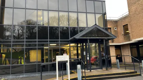BBC The entrance to West Cumbria Magistrates' Court. There is a small white sign indicating the entrance, where two steps lead to the front door. The front of the building is all glass, with black details surrounding individual panes.