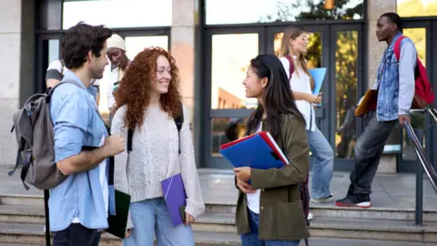 A group of students stand chatting on steps outside a campus building, carrying backpacks, notebooks and folders, while others walk past the entrance behind them.
