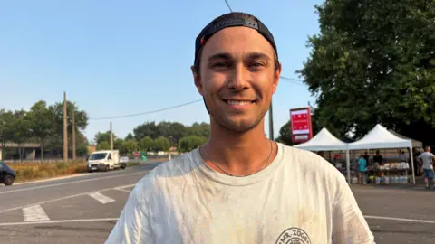 A man wearing a black baseball cap backwards on his head and a white t-shirt grins at the camera as he stands near a road