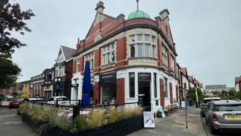 Joshua Rimmer a shot of the cafe from the outside. There is a seating area and black door. The cafe is located on a corner. 