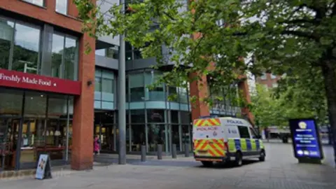 A police van is parked on a pedestrian walkway area where there is a digital sign, bollards and a glass and red brick fronted building