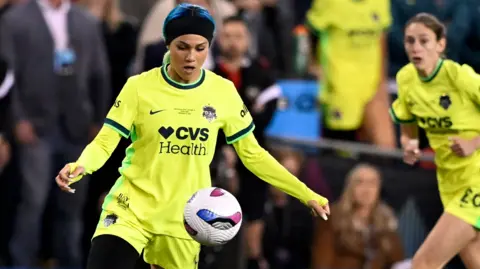 Trinity Rodman, in a neon yellow football kit, plays the ball during a game. A teammate watches in the background.